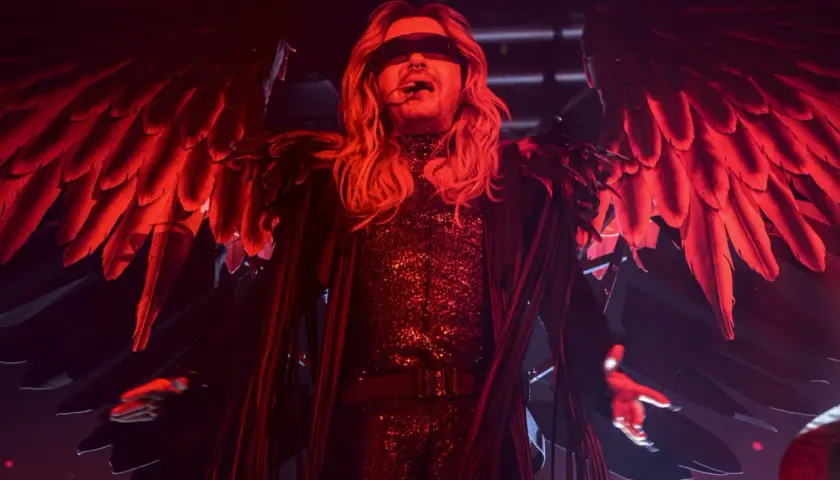 Bill Kaulitz z Tokio Hotel na koncercie w anielskich skrzydłach i błyszczącym stroju, w czerwonym świetle.