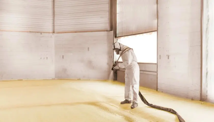 Un ouvrier en combinaison projette de la mousse polyuréthane sur le sol d'un entrepôt pour une isolation efficace.