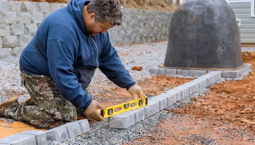 Mężczyzna klęczy, używając poziomicy do układania kostki brukowej na betonie. Powstaje ścieżka wokół pieca.