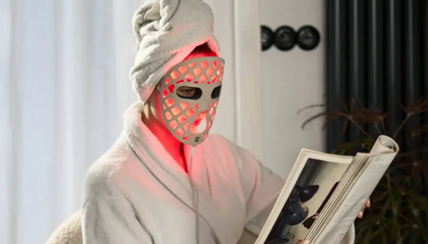 Person med handduk på huvudet och en led mask ansikte läser en bok i en morgonrock.
