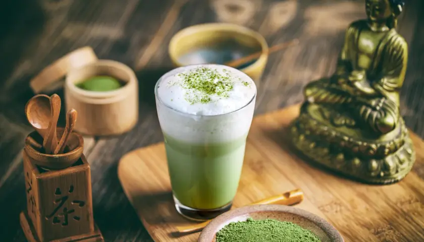 Szklanka zielonej herbaty matcha z pianką, obok naczynia z proszkiem matcha i bambusowe akcesoria.