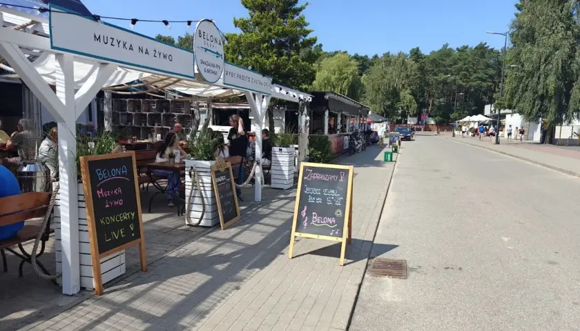 Dębki promenada: lokal Belona zaprasza na muzykę na żywo i koncerty. W tle widać letnie ogródki i spacerujących ludzi.