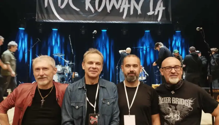 Czterech mężczyzn pozuje na tle sceny z banerem "ROCKOWANIA". Jeden z nich ma na sobie czerwoną kurtkę, inny dżinsową koszulę. W tle widać zespół i sprzęt. Idziemy do przodu dżem!
