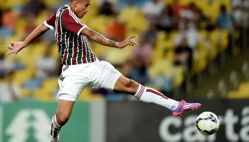 Piłkarz w koszulce Fluminense kopie piłkę na tle trybun.