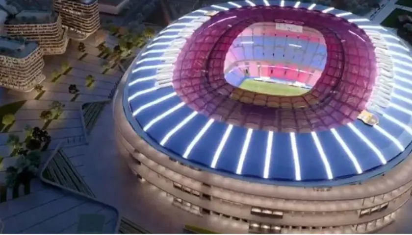 Nowe Camp Nou w nocnej odsłonie, z nowoczesną, oświetloną fasadą i dachem. Widać fragmenty otoczenia z budynkami i zielenią.