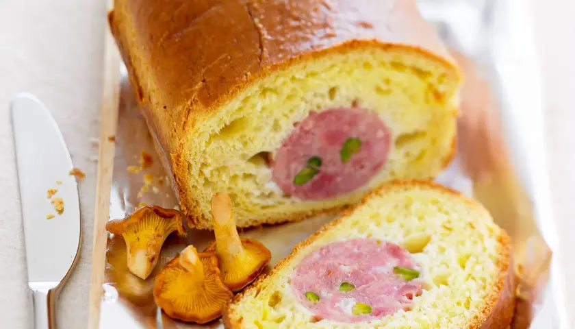 Un saucisson brioché doré, coupé en tranches, révèle sa garniture rosée. Des girolles et un couteau complètent cette gourmandise.