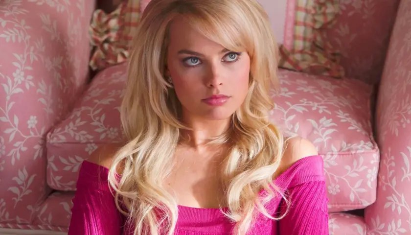 Margot Robbie, dans un film, assise sur un canapé rose à motifs floraux, porte un haut rose vif.