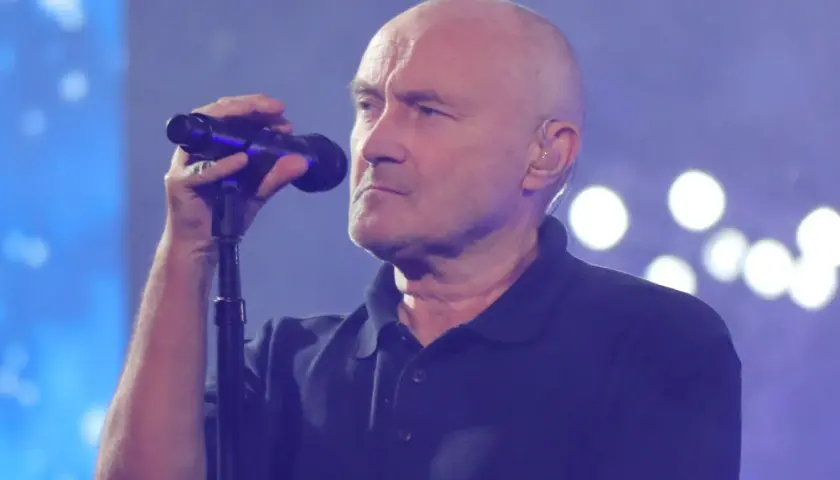 Phil Collins, wokalista zespołu Genesis, śpiewa do mikrofonu na tle niebieskiej sceny z białymi światłami.