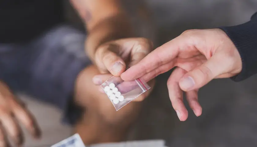 Dwie dłonie, jedna podaje, druga odbiera małą torebkę z białymi tabletkami, być może mdma.