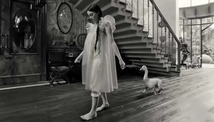 Bella Baxter, con un'aria sognante, cammina a piedi nudi in una casa d'epoca, accompagnata da un'oca e un cane.
