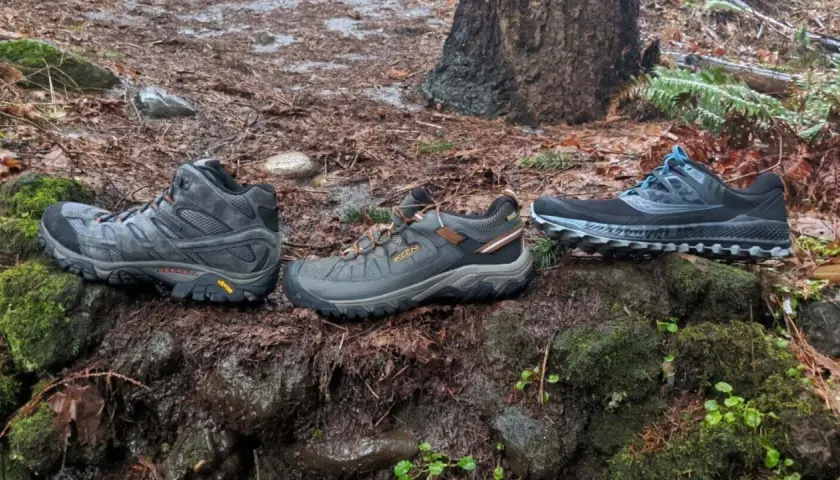 Three pairs of shoes sit on a mossy, wet forest floor: a mid-height hiking boot, a low-cut hiking shoe, and a pair of trail running shoes.