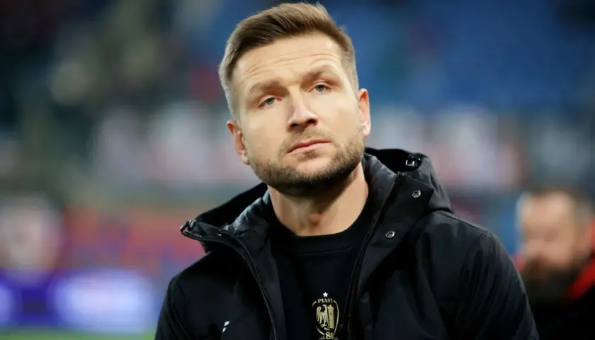 Mężczyzna w czarnej kurtce, z zarostem, w tle trybuny. To piast gliwice trener.