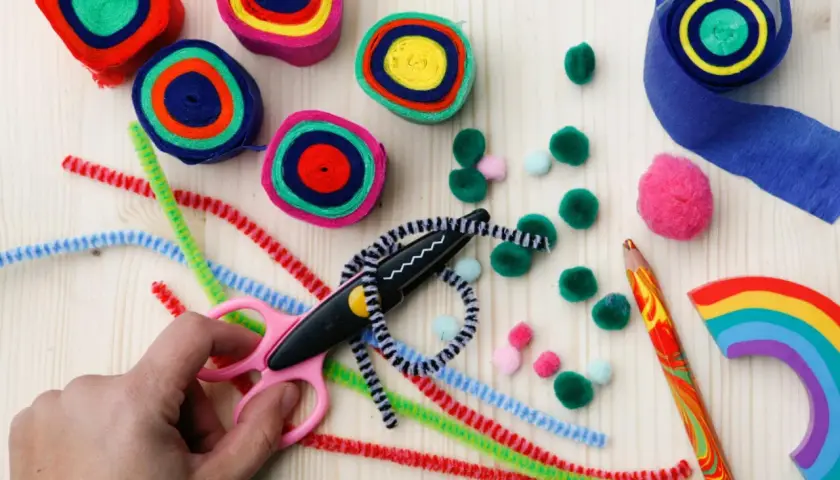 Bunte Rollen aus Papier, Pfeifenreiniger, Pompons und eine Schere laden zum Basteln ein. Ein Regenbogen-Holzstück und ein bunter Bleistift liegen bereit.