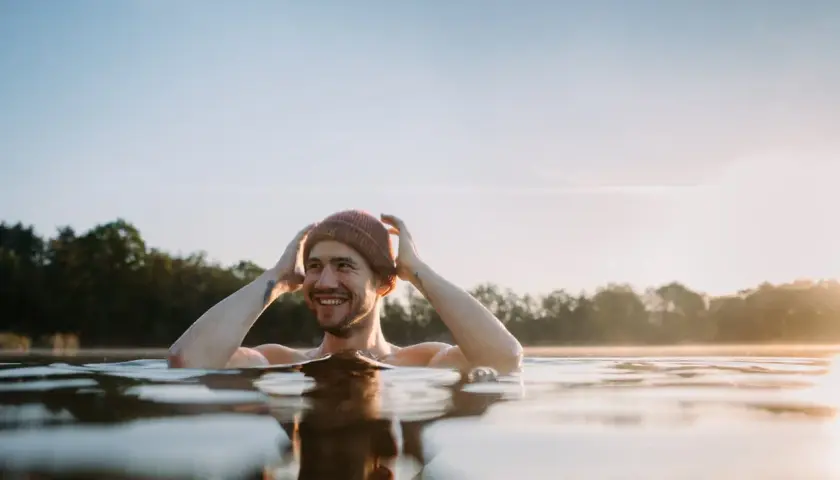 Uśmiechnięty mężczyzna w czapce zanurzony w zimnej wodzie, czerpiący radość z metody wim hof oddychanie.