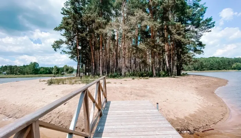 Drewniany pomost prowadzi do piaszczystej plaży otoczonej lasem sosen.