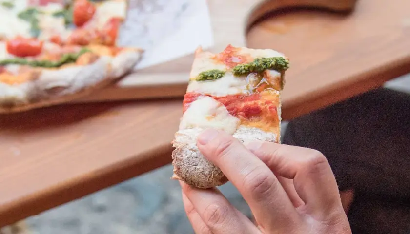 Eine Hand hält ein Stück Pinsa mit Tomaten, Mozzarella und Basilikumpesto. Was ist Pinsa? Eine römische Spezialität, die sich von Pizza unterscheidet.