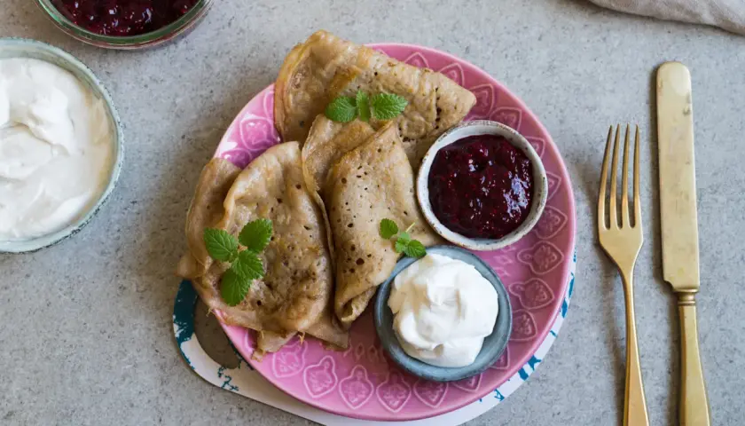 Glutenfria pannkakor med sylt och grädde på en rosa tallrik. En gaffel och kniv ligger bredvid.