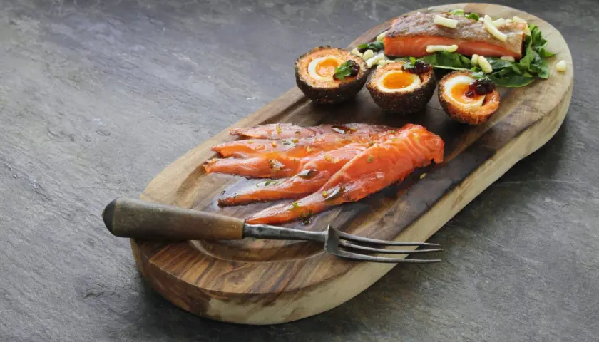 Tranché de truite fumée, œufs panés et garniture sur planche en bois. La cuisson truite fumée est mise en valeur.