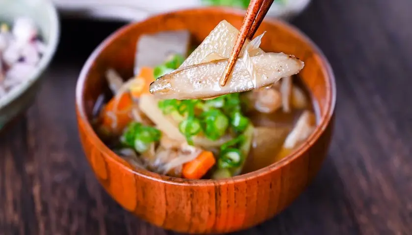 Chopsticks lift a piece of daikon from a steaming bowl of Japanese vegetable soup, filled with carrots, mushrooms, and green onions.