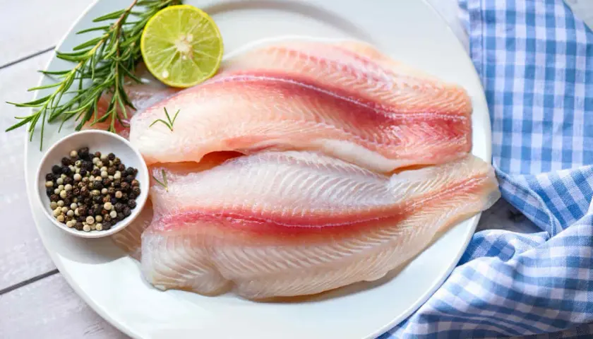Dos filetes de tilapia frescos, listos para cocinar, acompañados de romero, limón y granos de pimienta.