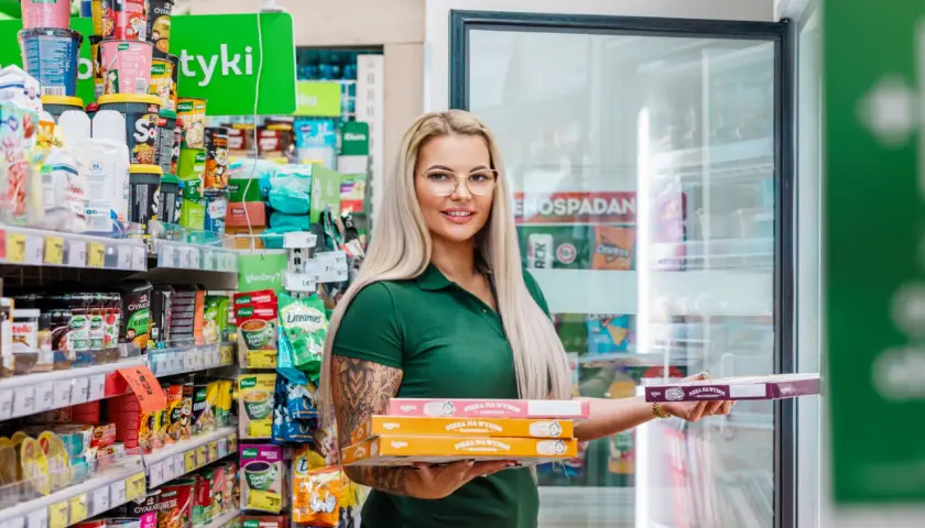 Kobieta w zielonej koszulce trzyma stos pudełek z pizzą na wynos w sklepie osiedlowym.