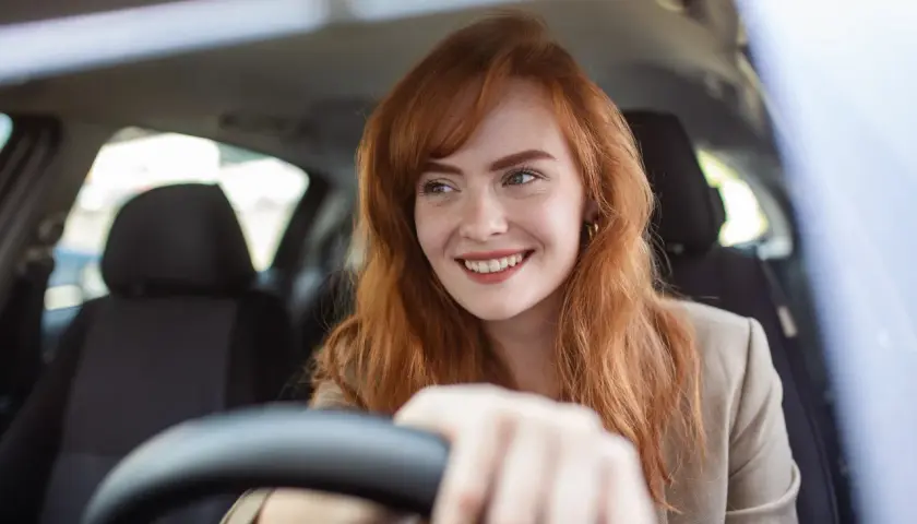 Donna sorridente al volante, con capelli rossi e lentiggini. Pensa alla sua assicurazione giornaliera auto per un viaggio sereno.