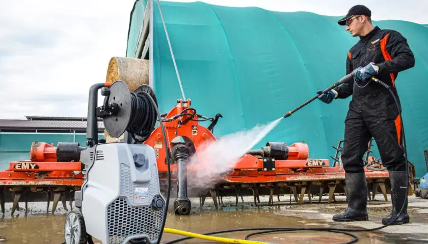 Hombre con traje de trabajo y botas limpiando maquinaria agrícola con hidrolimpiadora. Aprende como usar hidrolimpiadora para dejar todo impecable.
