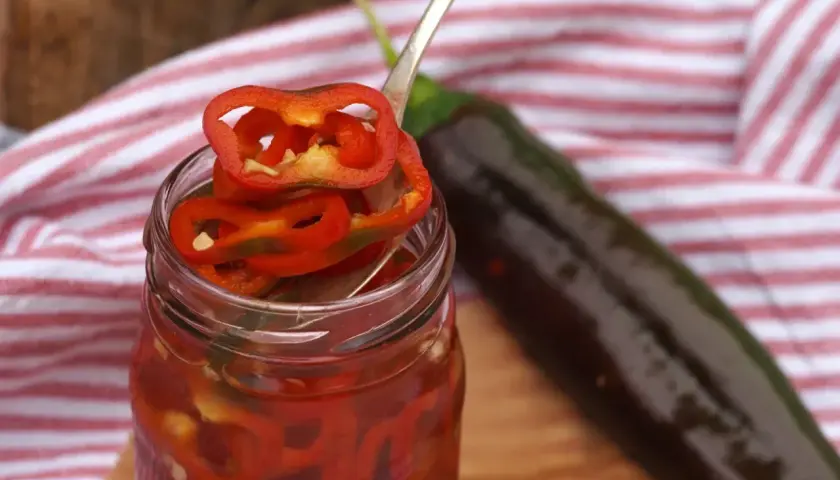 Burk med picklad chili, en sked med chiliringar och en hel mörk chili i bakgrunden.