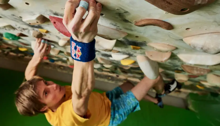 Alex Megos klettert konzentriert an einer Boulderwand. Seine Hand greift fest einen Griff, während sein Blick nach oben gerichtet ist.