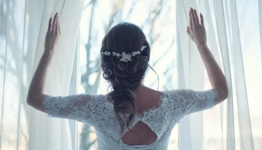 Elegancka fryzura dla świadkowej na wesele, z delikatną ozdobą we włosach, w białej koronkowej sukni, odsłaniająca zasłony.