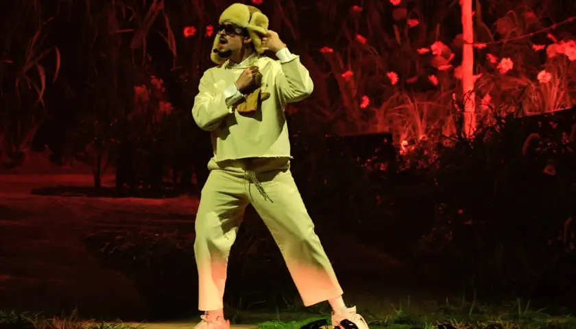 Bad Bunny w beżowym dresie i futrzanej czapce, jakby z "bad bunny café con ron tekst", tańczy na tle czerwonych kwiatów.