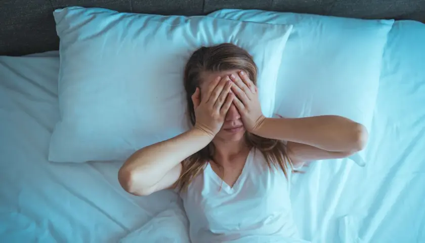 Mujer en la cama, con las manos cubriendo sus ojos. Son las 4 de la mañana y no puedo dormir, la luz azulada ilumina la escena.
