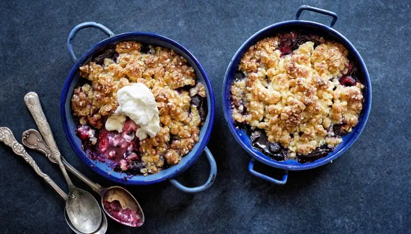 Två blåa ugnsformar med glutenfri smulpaj fylld med bär och toppad med grädde. Serveras med antika skedar.