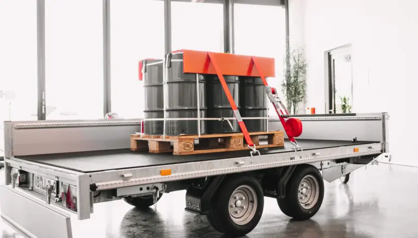 Fass auf einem Anhänger sind drei Fässer auf einer Palette sicher mit roten Gurten und einem orangefarbenen Halter frei verladen.