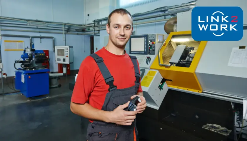 Uśmiechnięty tokarz w warsztacie, trzymający przyrząd pomiarowy, obok nowoczesnej maszyny CNC.