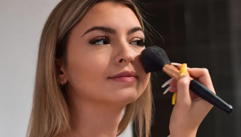Młoda dziewczyna z długimi blond włosami nakłada puder pędzlem. Jej makijaż jest subtelny, podkreślający naturalne piękno, w stylu "clean girl".