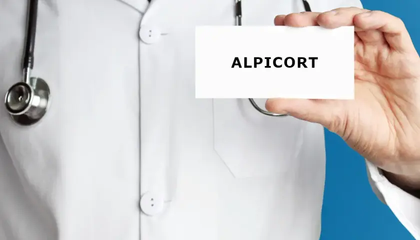 Lekarz w białym kitlu prezentuje kartkę z napisem ALPICORT. Stetoskop wisi na jego szyi.