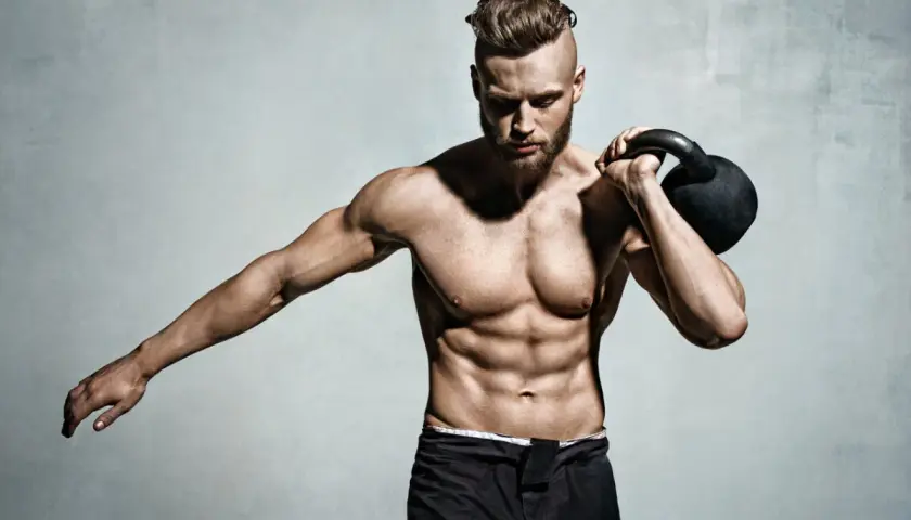Mężczyzna z wyrzeźbionym brzuchem i kettlebell na ramieniu. Pokazuje, jak wejść na masę, trenując siłowo.