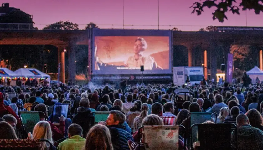 Stor publik njuter av sommarbio Stockholm under en lila kvällshimmel. En film visas på en stor duk framför tält och träd.