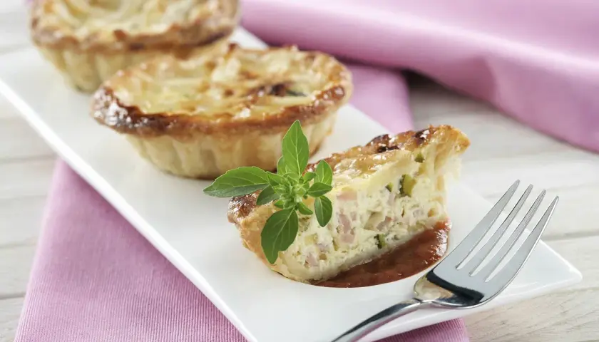 Trois mini quiches sans pâte garnies d'une tranche coupée, sur une assiette blanche avec une feuille de basilic.