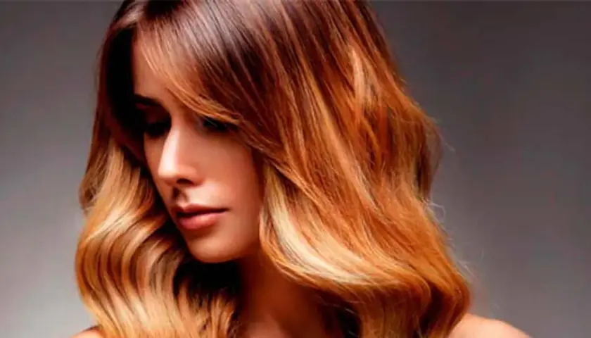 Mujer con pelo californianas, con ondas y un degradado de color castaño a rubio dorado, luciendo un estilo moderno y radiante.