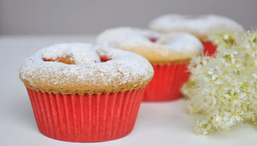 Pyszne muffinki dla dzieci, posypane cukrem pudrem, w czerwonych papilotkach, obok delikatnych białych kwiatów.
