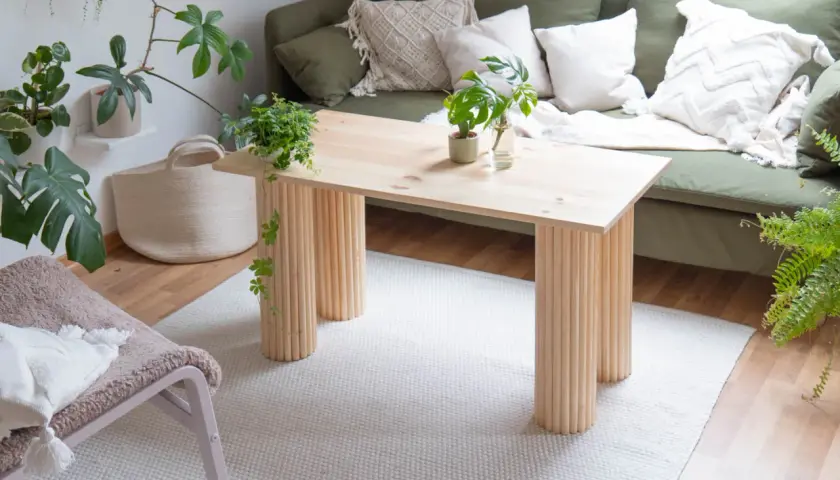 Ein heller Holztisch mit säulenartigen Beinen, perfekt für ein DIY-Projekt. Daneben ein grünes Sofa, Pflanzen und ein Körbchen.