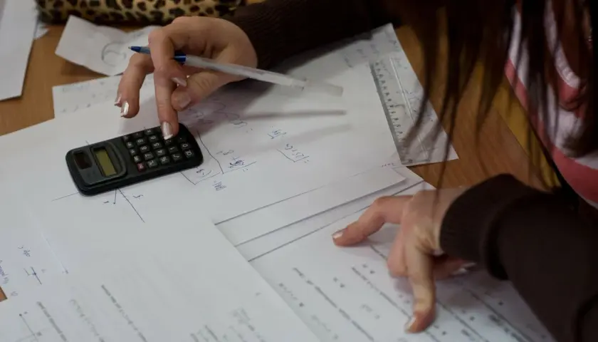 Dłoń ucznia z kalkulatorem i długopisem nad kartkami z matematycznymi obliczeniami.