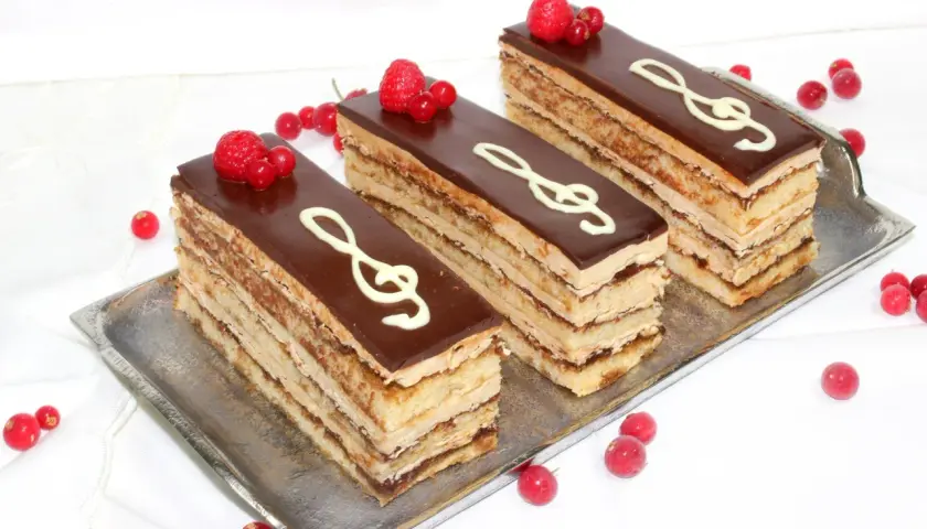 Tres porciones de pastel ópera, con capas de bizcocho y crema, cubiertas de chocolate y decoradas con clave de sol y frutos rojos.