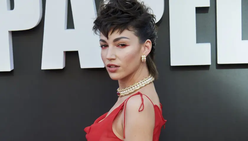 Una mujer con un peinado mullet chico y maquillaje llamativo posa con un vestido rojo y joyas doradas.