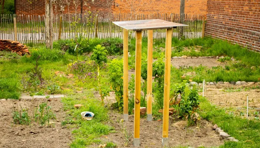 Ein provisorisches tomatendach selber bauen: Holzpfosten mit Metallfüßen tragen eine einfache Dachkonstruktion im Garten.