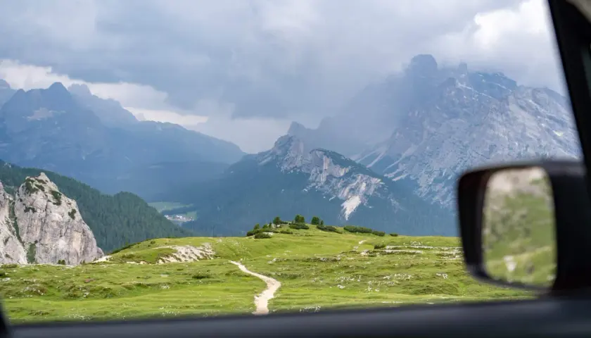 Vue depuis une voiture en Italie, traversant des prairies verdoyantes et des montagnes majestueuses sous un ciel nuageux.