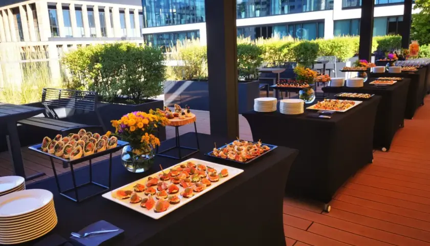 Elegancki catering Warszawa na imprezę plenerową. Stoły z przekąskami i napojami na tarasie z widokiem na nowoczesne budynki.
