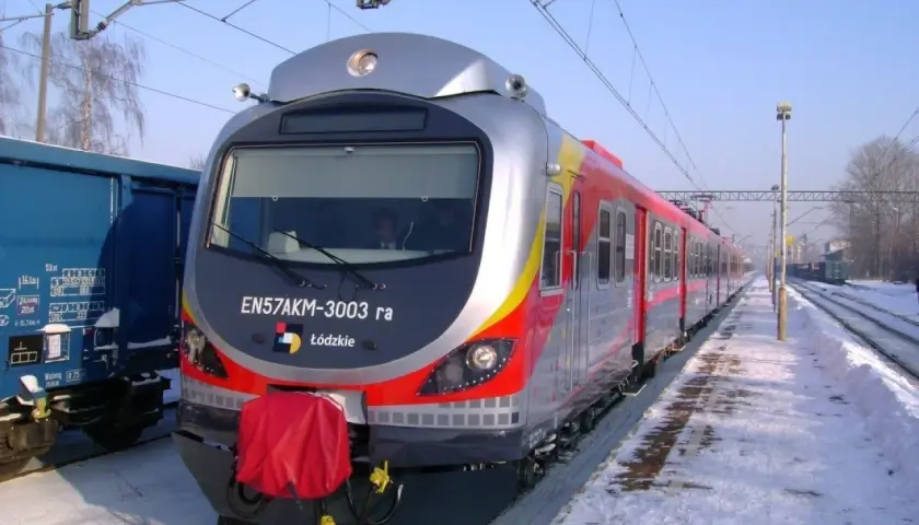 Pociąg EN57AKM-3003 ra z Łodzi stoi na zaśnieżonym peronie. Obok niebieski wagon towarowy.